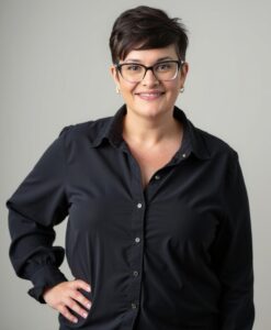 Website 1 White woman wearing a black button down shirt. She is standing with her hand on her hip. She has dark hair cut into a pixie and wear black cat-eyed glasses.